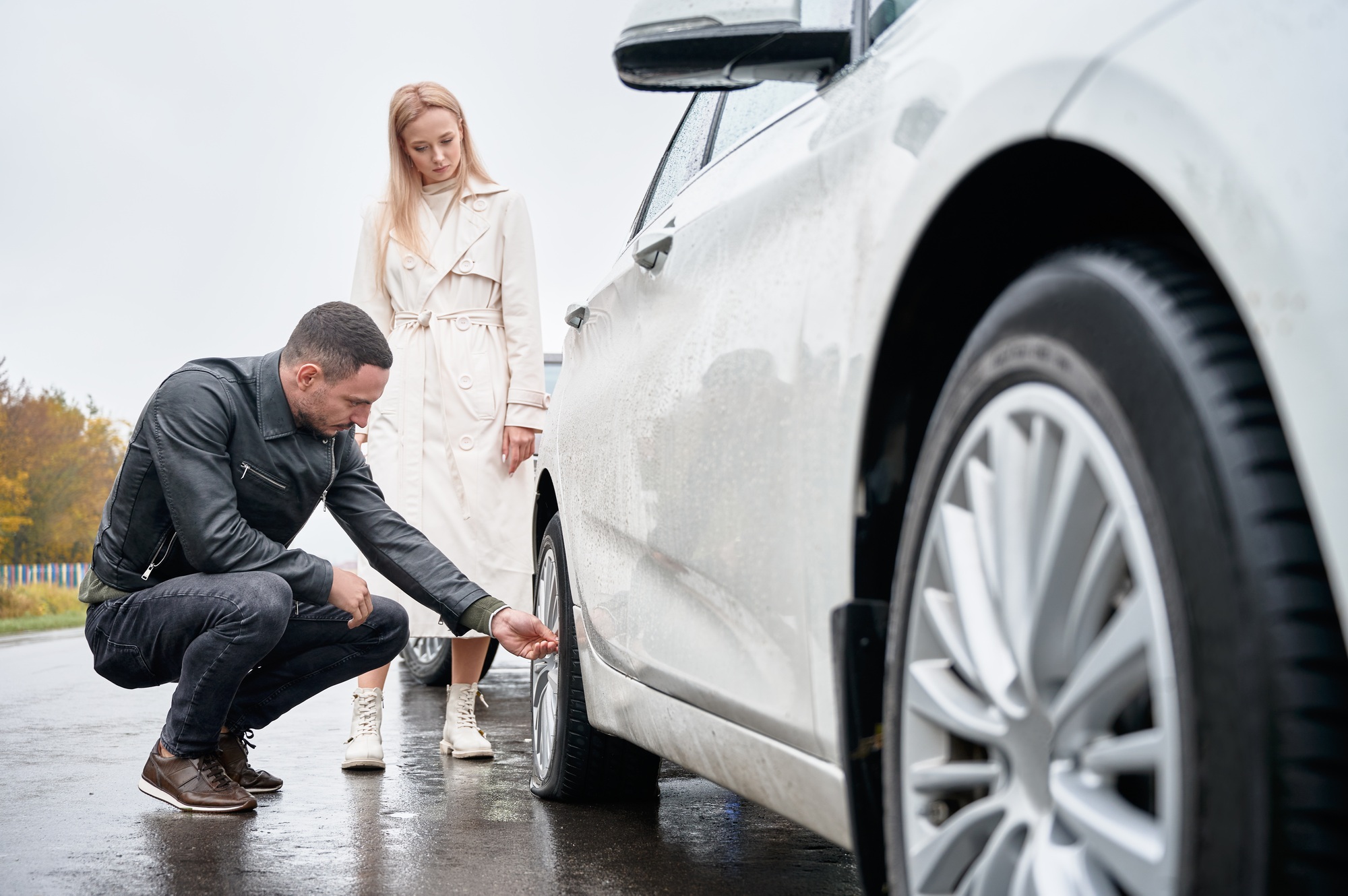 Man inspecting damaged tire of girl's white car.